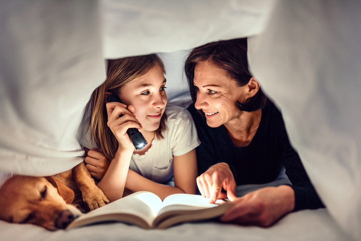 Mother and daughter with their dog looking at a book with a flashlight under sheets.
