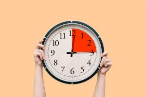 An image of a face of a clock, held up by two hands in front of an orange background.