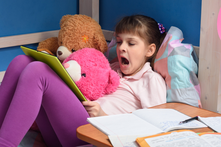 Young girl with ADHD, yawning in bed, doing school work with her stuffed animals for at-home learning