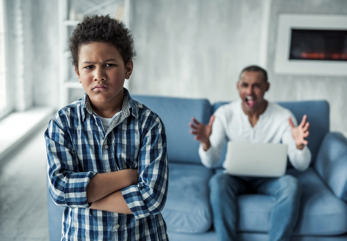 Young boy with ADHD and anger crossing his arms and turning his back to his father who is looking frustrated at him and sitting on the couch with his laptop