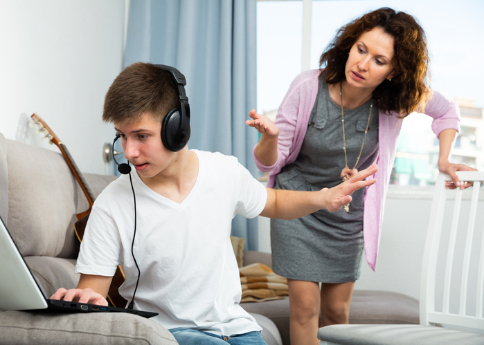 Mother trying to get son's attention who is on the computer listening to headphones and raising his hand for his mom to go away