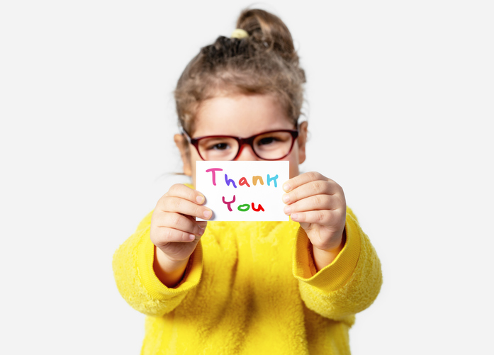 Toddler in a yellow shirt holding up a handwritten card that says "Thank you" in different colors.