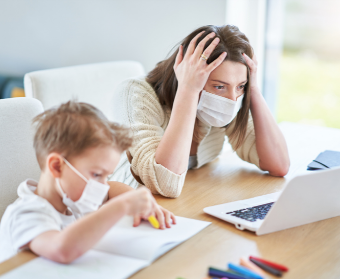 Mother looking overwhelmed as she holds her hand on her head looking at a computer with a mask on next to her child who is doing remote work during the pandemic