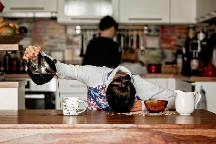 A person pouring coffee onto the table, missing the cup, with her head down on the kitchen table.