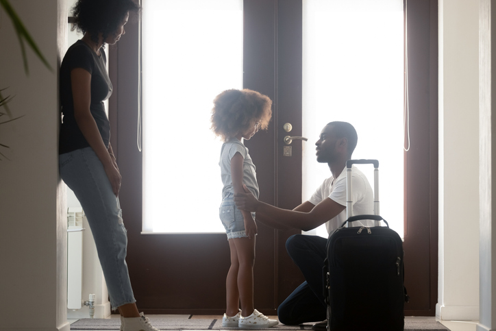 Child with ADHD and her divorced parents, standing as she says goodbye to her father who is kneeling infront of her with his suitcase by the door, while her mother stands by and watches