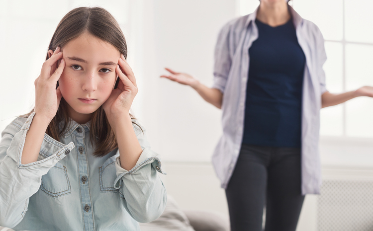 An adolescent with ADHD holds her hands to her temples as she looks at the camera, annoyed as her mother holds their hands out in question in the background.