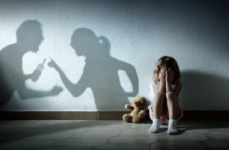 Child with ADHD sitting on the floor with her teddy bear in the dark with her eyes over her head as shadows of her divorced parents fighting are on the wall