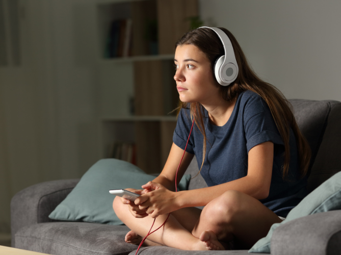 Teen with ADHD looking out the window while sitting on the couch wearing overear headphones plugged into the phone