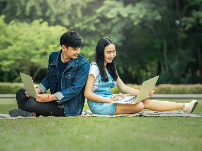 Two teens having a good time sitting on a grass and doing computer work.
