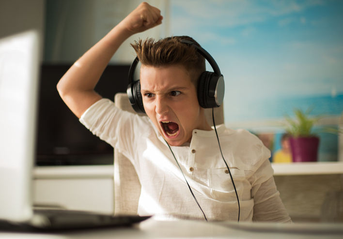 Boy with ADHD wearing headphones looking upset raising a fist above his head as he looks at his laptop