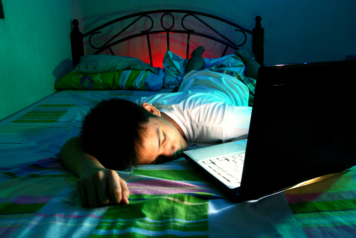 Boy with ADHD sleeping on the wrong side of his bed at night next to his laptop which is open and on