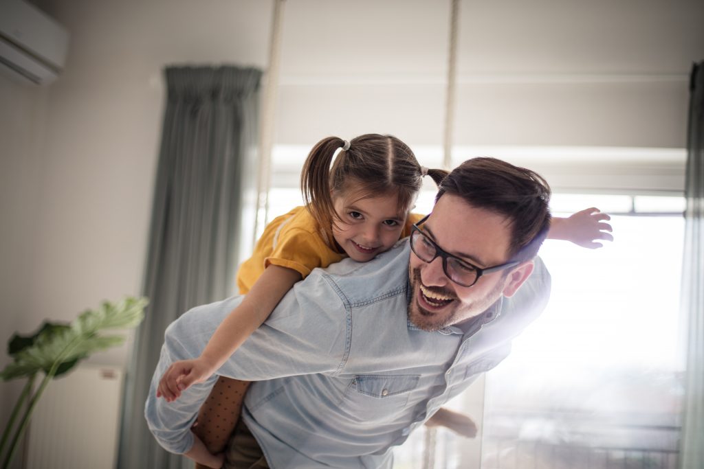 Father in a light blue long-sleeve collared shirt laughs and bends over as he walks around his living room holding his daughter with pigtails and a yellow shirt on his back. She has her arms out like she's an airplane, and their both laughing and having fun.