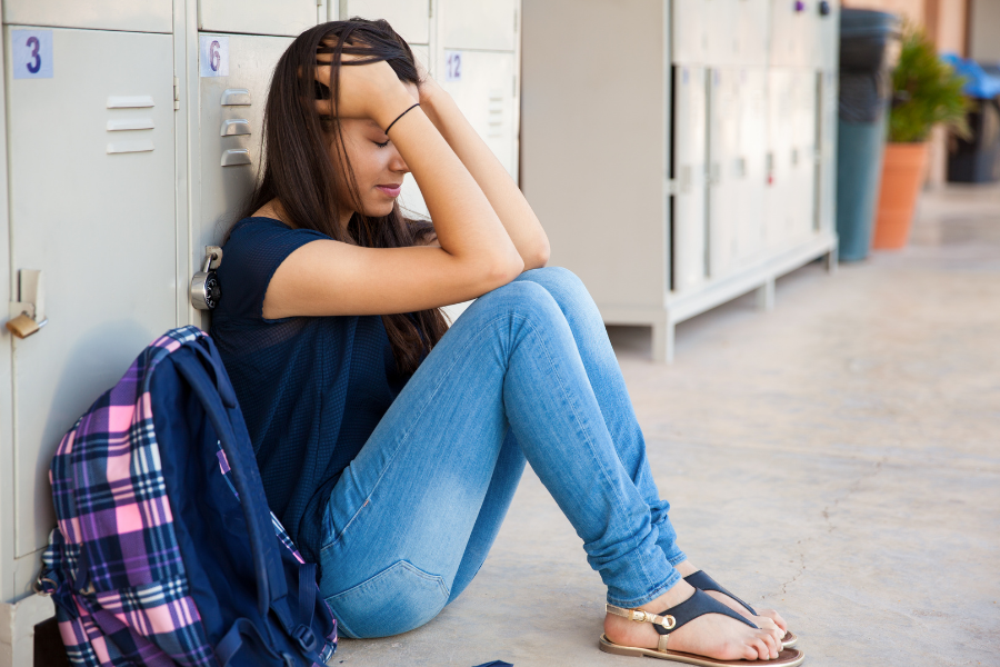 Teen girl with ADHD sitting on the ground in front of her locker with her hands on her head looking anxious and down