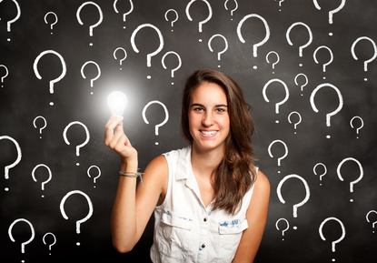 A neurodiverse teen girl holding a lit lightbulb in-front of a black background that has big white question marks all over it.