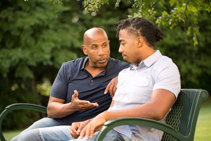 A father practicing parental scaffolding strategies and having an important discussion with his neurodiverse teen outside on a bench under a tree