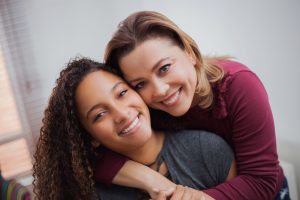 A mother hugging her neurodiverse adolescent daughter around the shoulders from behind while they both smile and look at the camera