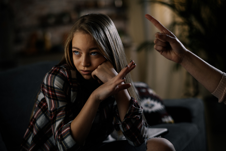 Girl teen looking annoyed as she sits on the couch while her parent responds to her ADHD and negativity by pointing their finger at her.