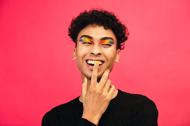 Neurodiverse and gender-diverse teen wearing rainbow eye makeup and happy face nail polish laughing and looking to the side in front of a pink background
