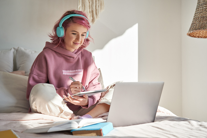 Neurodivergent teen with ADHD with pink sweatshirt and pink hair and teal headphones on a bed doing homework with on a laptop in her room