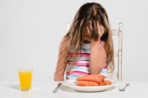 Girl with ADHD looking down with her palm on her face in front of her dinner plate that has a pile of carrots beside a cup of orange juice