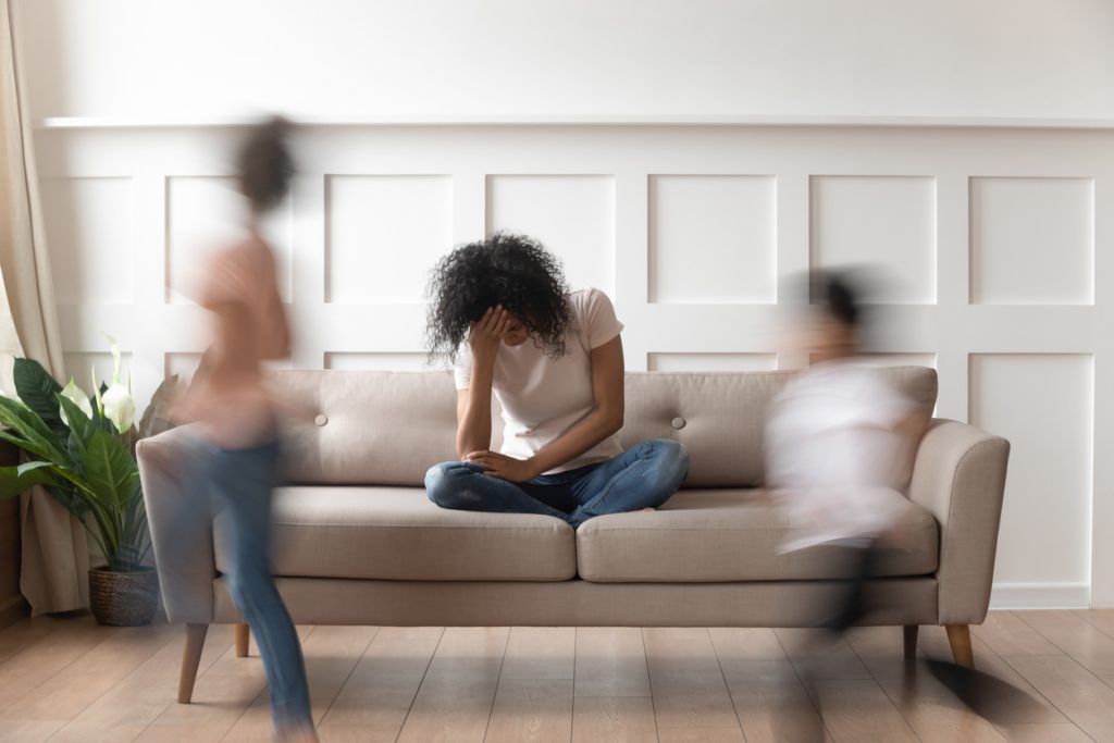 Mother sitting on the couch with her head on her hands while her kids run past out of focus.