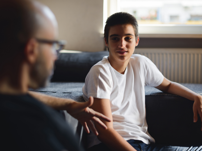 Neurodiverse teen boy and father making eye contact and having a conversation in the living room, view point from behind father.