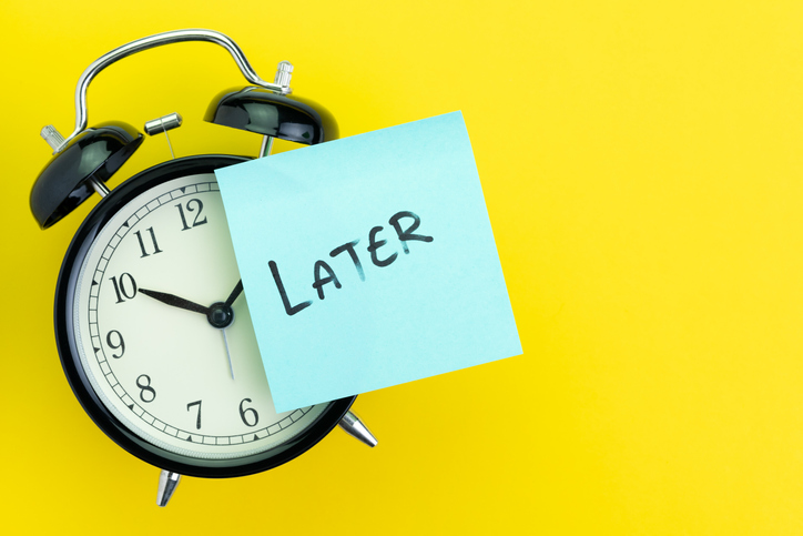 A black alarm clock in front of a bright yellow background with a teal sticky note that says "LATER" on the clock.