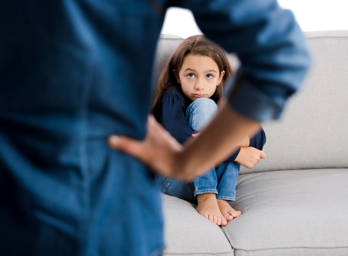 Young girl with ADHD curled up looking scared about getting a time-out on the couch, while viewing an upset parent with their hand on their hip looking at her