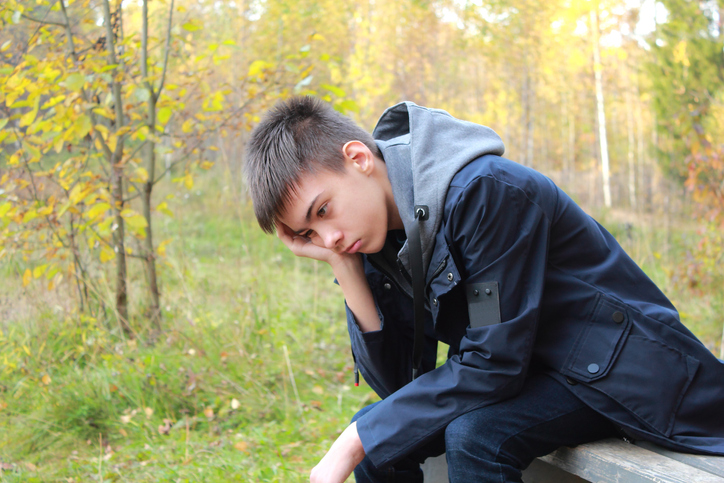 Teen boy sitting outside by trees, looking sad and down at the ground with his head resting down on his hand.