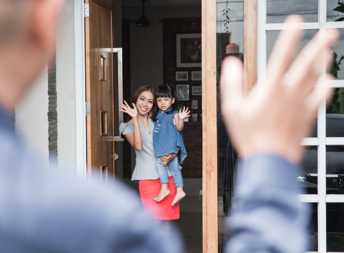 A mother holding her young daughter in her house waving to someone outside