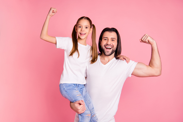Father holding up his tween daughter while they both smile and flex their muscles in front of a pink background.