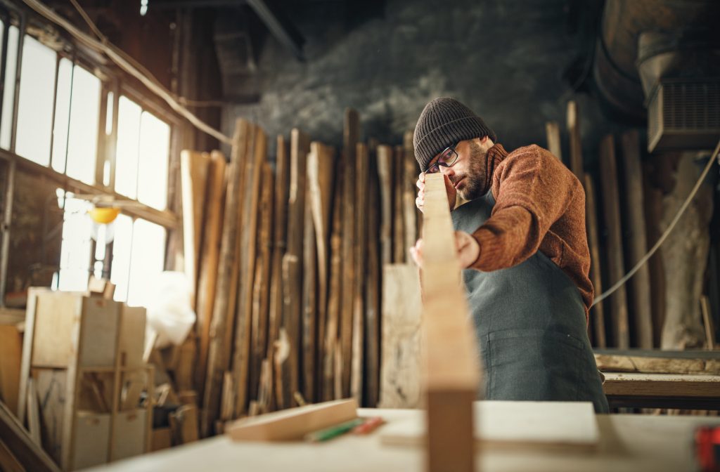 Focused adult male woodworker in apron and knitted gat cutting wooden plank while working in carpentry workshop