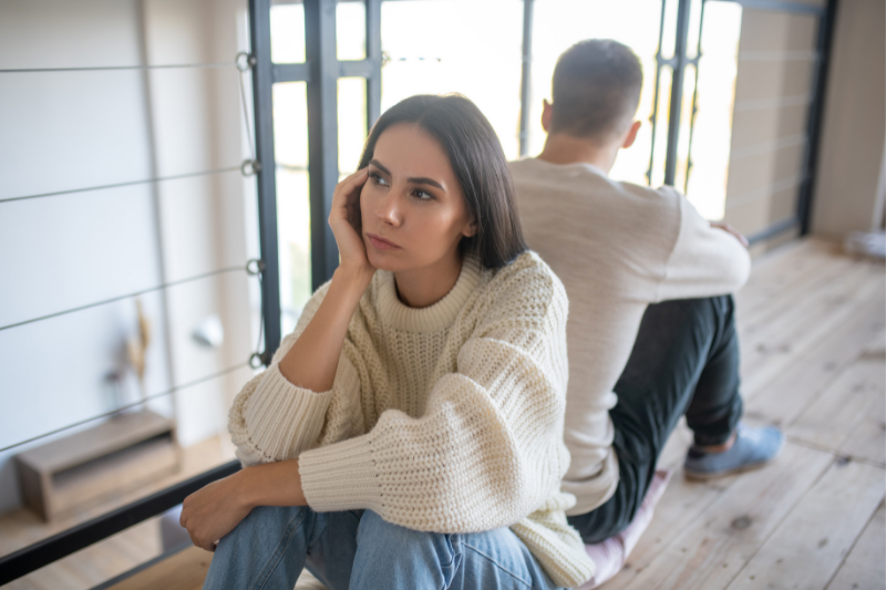 woman sitting on the ground back to back with her partner, looking upset but thinking
