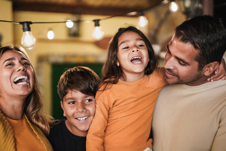 Family of four laughing, singing and hugging indoors under string lights.
