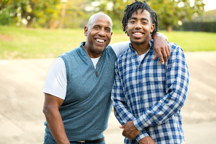 Father with his arm wrapped around his son, smiling together, standing outside at the camera
