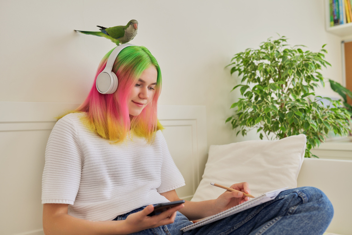 Girl with colorful hair sitting at home doing homework with a bird standing on her headphone on her head.