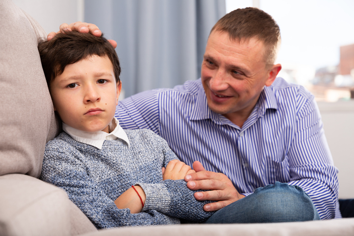 Young boy looking upset as his father sits next to him, trying to help him feel better