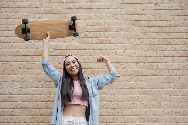 Teenage girl standing in front of a brick wall holding up a skateboard
