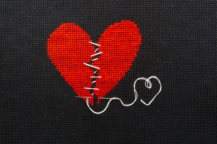 A stitching of a red broken heart being stitched together with white thread against a gray background.