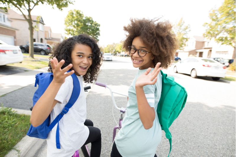 two girls on bikes with backpacks smiling and waving