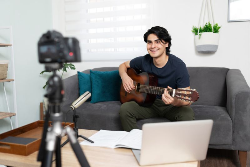 young man with guitar on couch smiling at camera on tripod